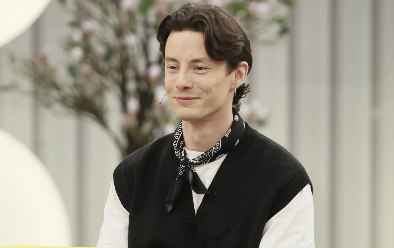 Andrzej Kliś, uczestnik programu "Farma" Andrzej Kliś, uczestnik "Farmy", w białej koszuli i czarnym swetrze, siedzi na tle rozmytego drzewa.