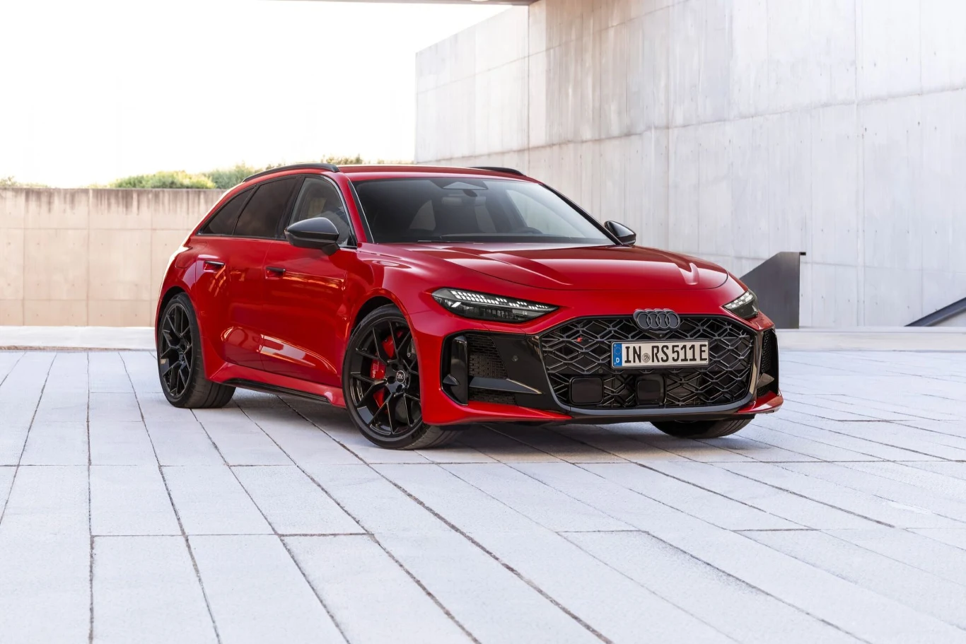 czerwone Audi RS na czarnych felgach stoi na nowoczesnej, jasnej nawierzchni obok betonowych ścian, podkreślając sportowy charakter auta dzięki wyrazistym liniom nadwozia i agresywnym wlotom powietrza w przednim zderzaku