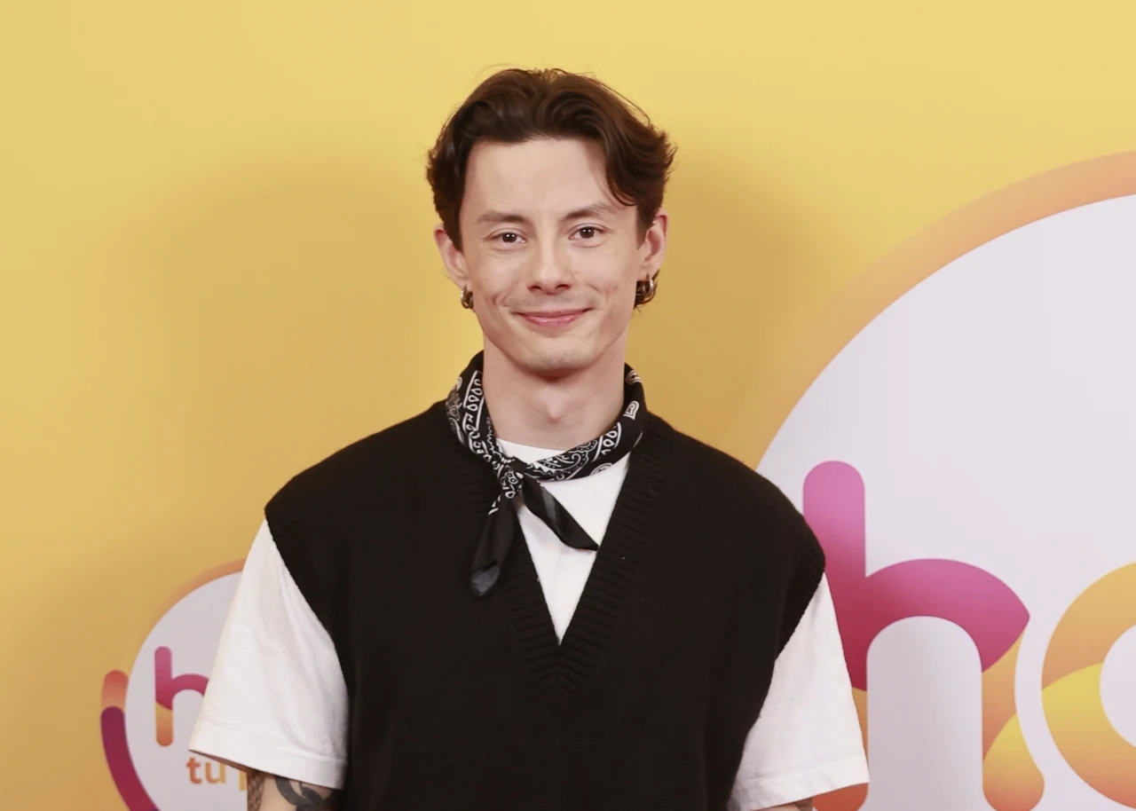 Andrzej Kliś w "halo tu polsat" Młody mężczyzna z krótkimi, ciemnymi włosami ubrany w czarną kamizelkę na białym t-shircie, z czarną bandaną zawiązaną na szyi, stoi na tle żółtej ścianki z elementami graficznymi i napisem w stylu pop-art.