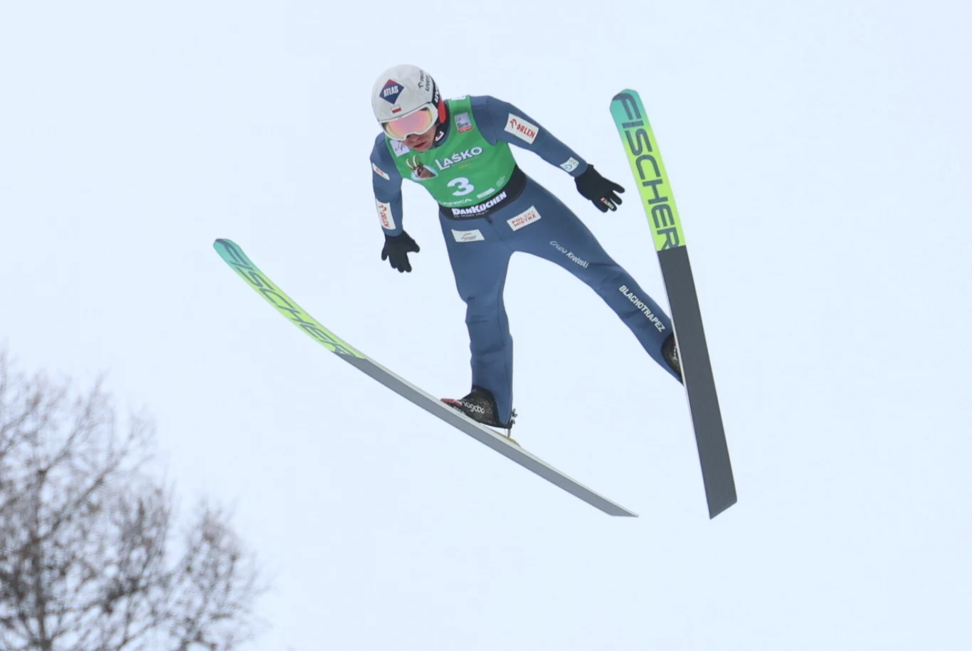 Skoczek narciarski w kombinezonie i kasku leci w powietrzu podczas skoku z wyciągniętymi nartami, widoczne logo Fischer na jednej z nart oraz numer startowy 3 na zielonej kamizelce.