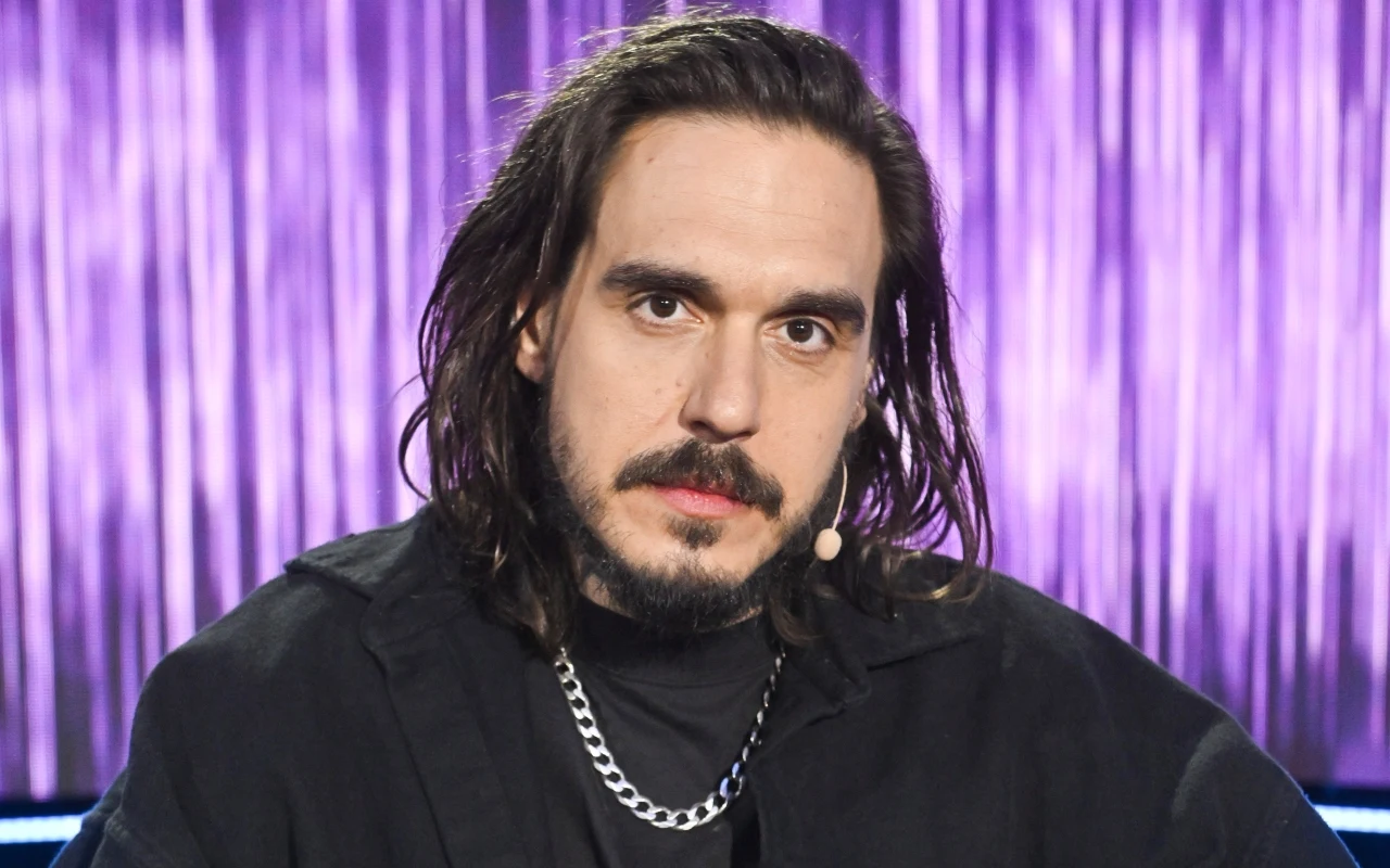 Miuosh Miuosh z długimi ciemnymi włosami i brodą, w czarnej koszuli, srebrny łańcuch, mikrofon przy twarzy, fioletowa kurtyna