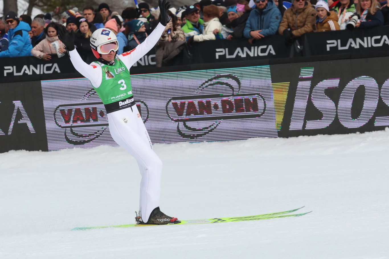 Skoczek narciarski w białym kombinezonie i zielonym plastronie z numerem 3 ląduje z rozłożonymi rękami na śniegu podczas zawodów, w tle widoczny tłum kibiców oraz banery reklamowe.