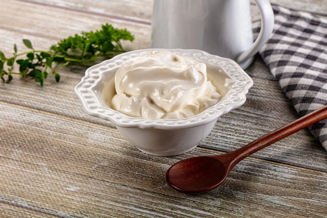 porcelanowa miseczka wypełniona kremową śmietaną, obok drewniana łyżka, w tle dzbanek oraz ściereczka w kratkę