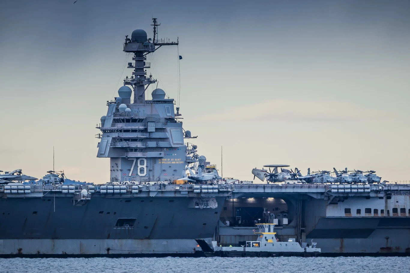 lotniskowiec wojskowy z numerem 78 na burcie, z wieloma samolotami myśliwskimi zaparkowanymi na pokładzie oraz małą jednostką pływającą przy kadłubie na tle pochmurnego nieba