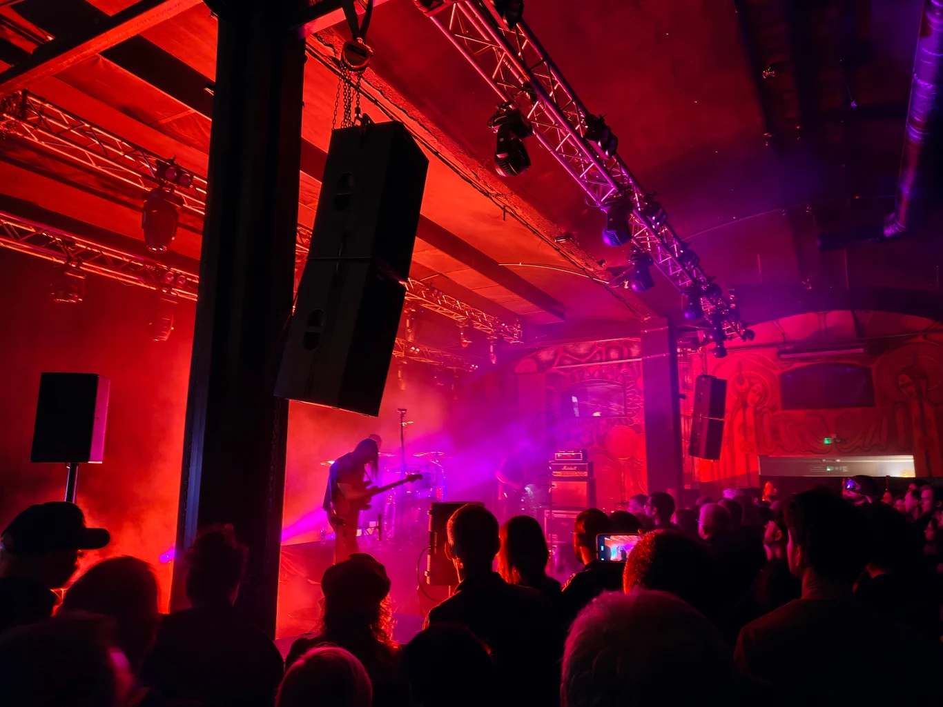 Koncert w klubie muzycznym z tłumem słuchaczy, kolorowe światła sceniczne oświetlają zespół grający na scenie, w tle widoczna ściana z dużym muralem.