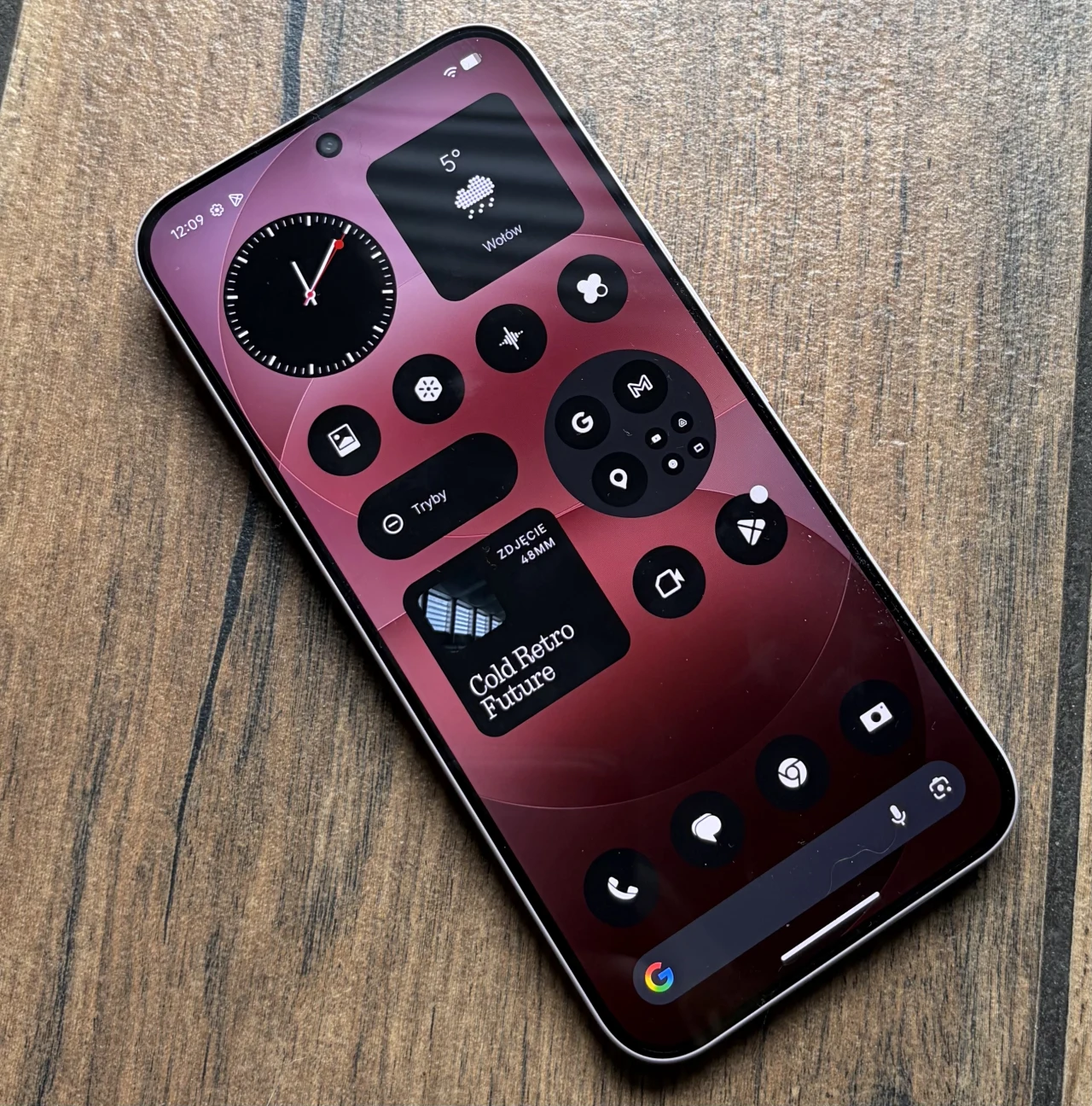 smartfon z ekranem wypełnionym czarnymi okrągłymi widgetami i ikonami na czerwonym tle, dominujące elementy to zegar analogowy, widżet pogody oraz panel skrótów, urządzenie leży na drewnianej powierzchni