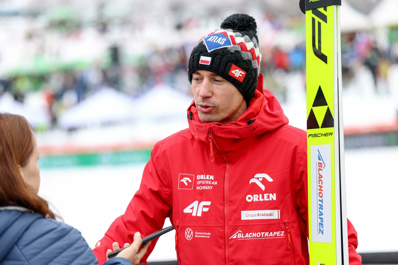 mężczyzna w czerwonej kurtce zimowej z licznymi logotypami partnerów udziela wywiadu dziennikarce, w ręku trzyma narty, na głowie ma czapkę z pomponem