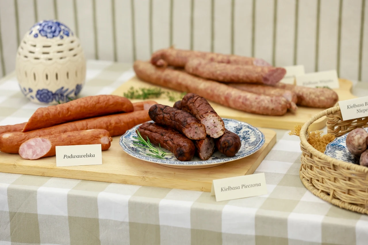 Kiełbasy na świąteczny stół Różne rodzaje kiełbas, w tym podwawelska i pieczona, ułożone na talerzach i drewnianych deskach na obrusie w kratę, z widocznymi etykietami opisującymi poszczególne produkty.