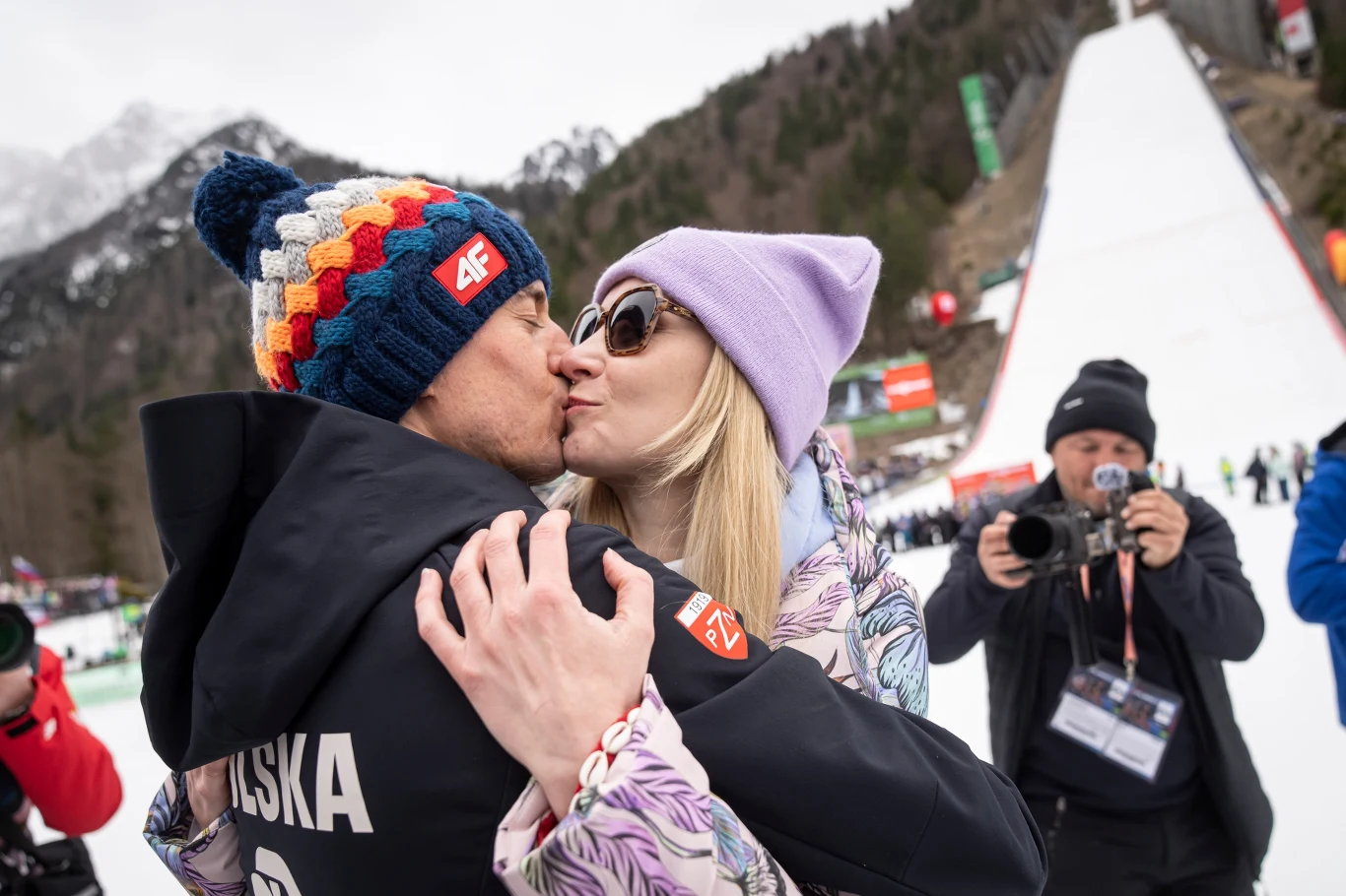 sportowiec w kurtce z napisem 'POLSKA' całuje kobietę w liliowej czapce i okularach przeciwsłonecznych, w tle fotografowie i skocznia narciarska otoczona górami