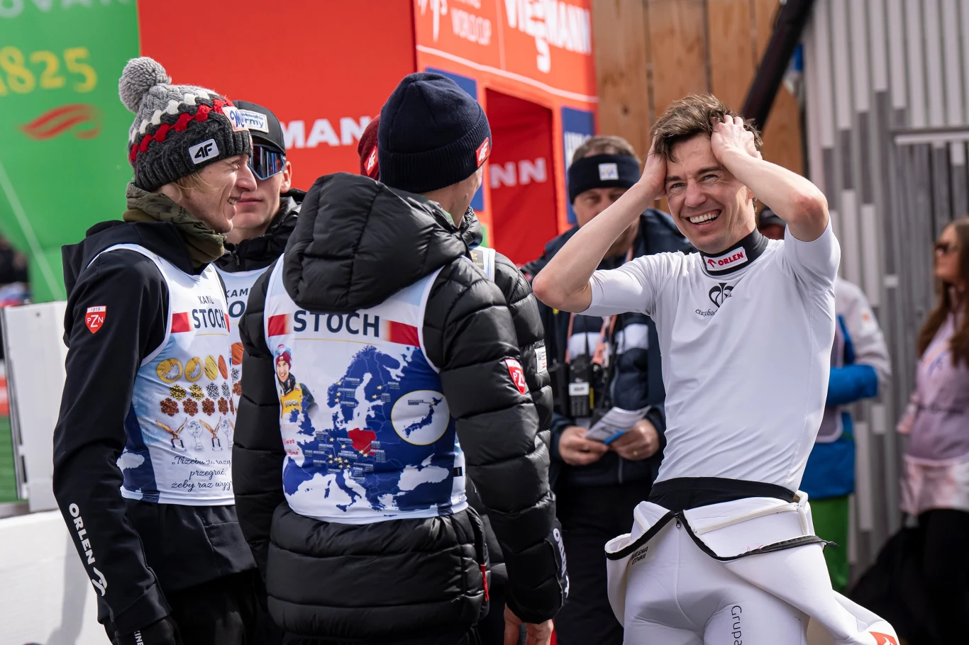 trzech mężczyzn w sportowych strojach rozmawia ze sobą na tle zimowej scenerii, jeden z nich trzyma ręce na głowie i uśmiecha się szeroko, panuje radosna atmosfera