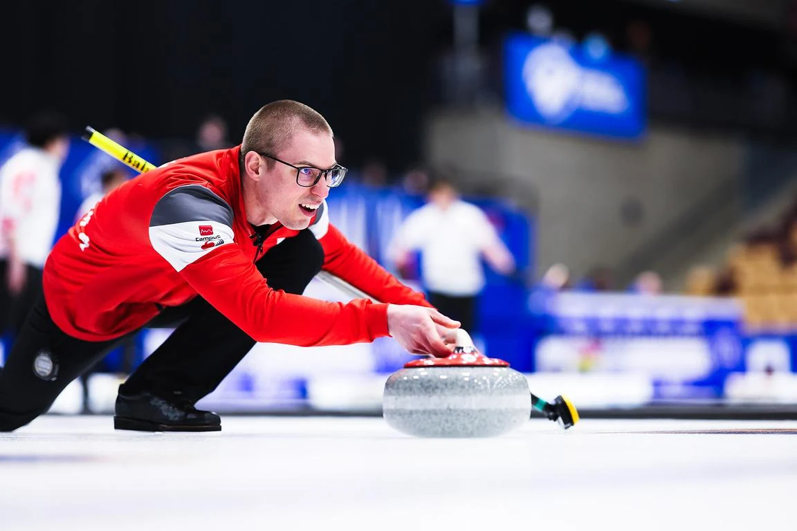 mężczyzna w czerwonej sportowej bluzie wypuszcza curlingowy kamień na lodowej tafli podczas rozgrywki, skoncentrowany wyraz twarzy, dynamiczna pozycja ciała