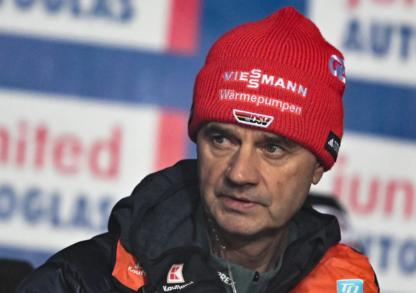 mężczyzna w czerwonej czapce z logo Viessmann i ciemnej kurtce zimowej patrzy w bok, na tle rozmytego banera z napisami