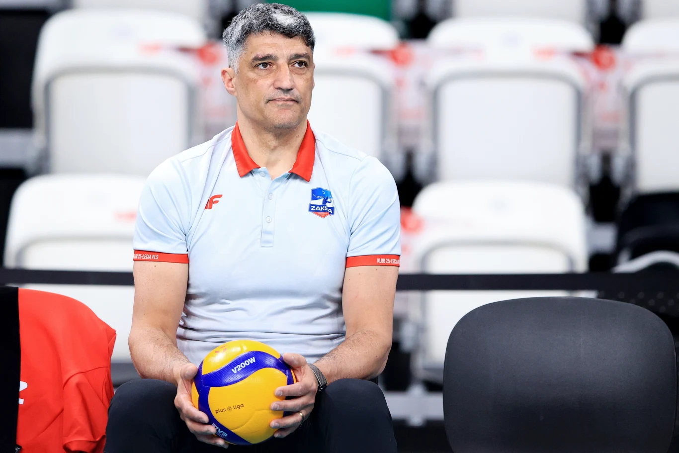 mężczyzna w jasnej koszulce z logo drużyny siatkarskiej siedzi na trybunach i trzyma w rękach piłkę do siatkówki, za nim puste krzesła