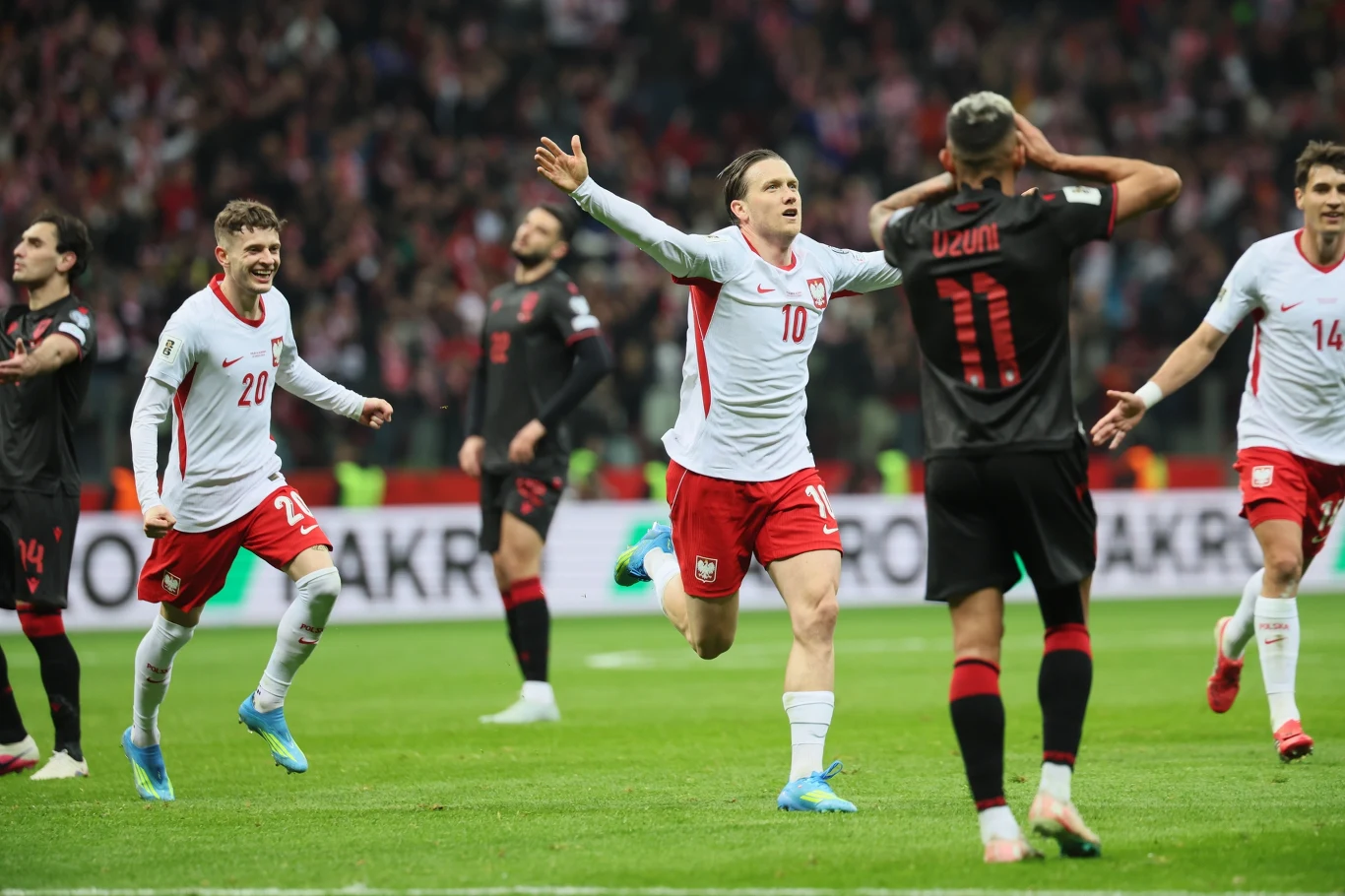 piłkarz w białej koszulce z numerem 10 unosi ręce w geście triumfu biegnąc po murawie, podczas gdy jego koledzy z drużyny również wyrażają radość; zawodnik drużyny przeciwnej z numerem 11 trzyma ręce na głowie w geście rozczarowania