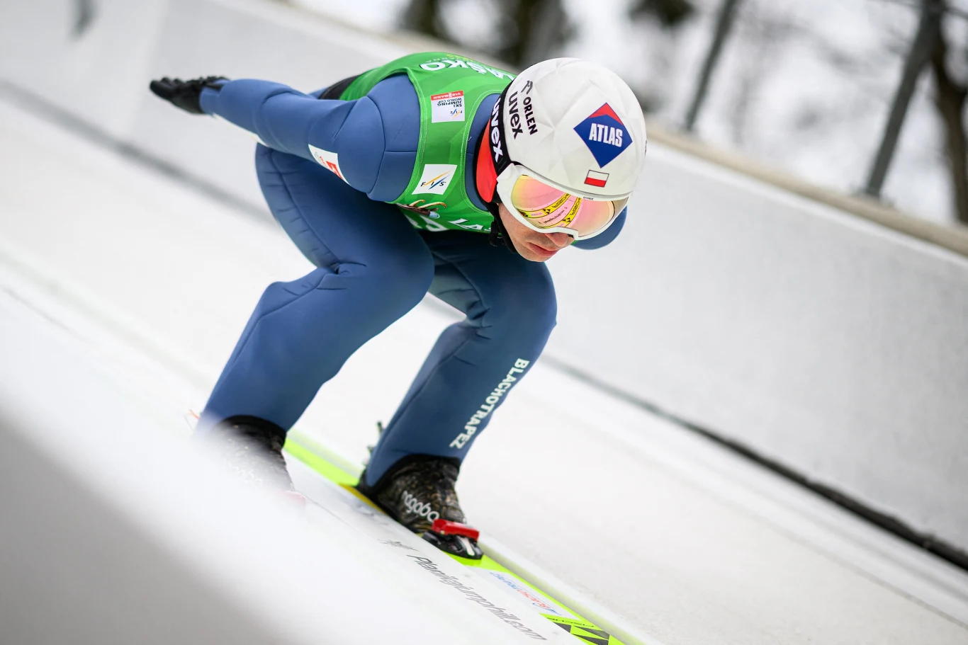 Skoczek narciarski w aerodynamicznej pozycji tuż przed wybiciem z progu, ubrany w kombinezon startowy, z kaskiem i goglami, jadący po torze rozbiegowym na nartach skokowych.