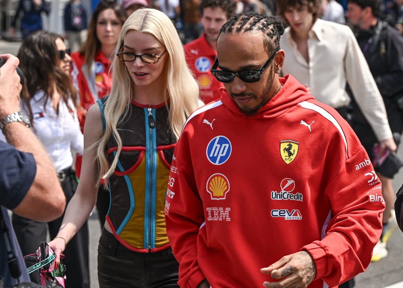 mężczyzna w czerwonej bluzie Ferrari z charakterystycznymi sponsorami i w ciemnych okularach idzie obok kobiety w okularach przeciwsłonecznych, ubranej w kolorową, designerską sukienkę; wokół nich przechadzają się inni uczestnicy wydarzenia, widoczne t...