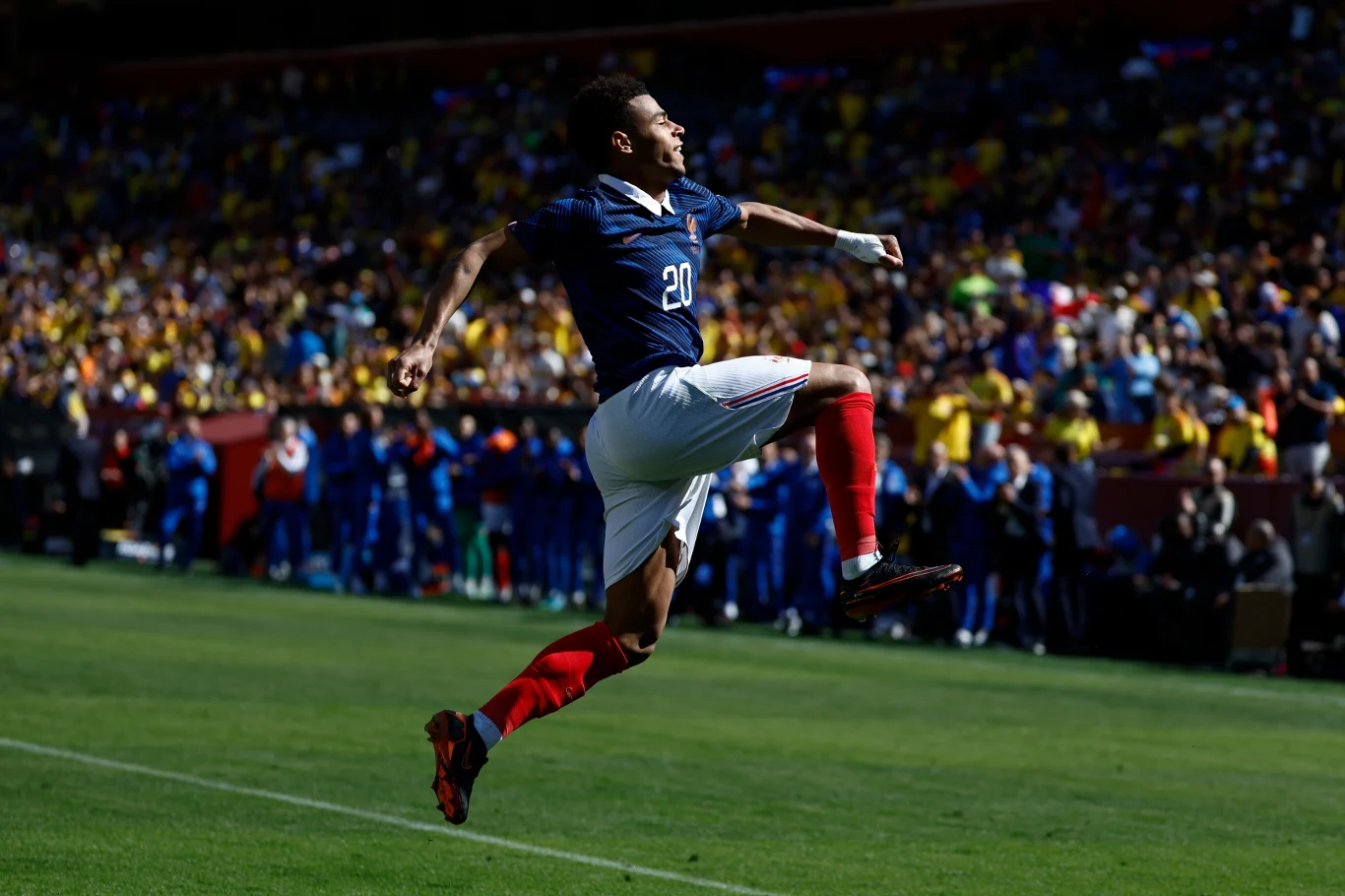 piłkarz w granatowej koszulce z numerem 20 wykonuje dynamiczny skok z uniesionymi ramionami na tle pełnych trybun w trakcie meczu piłki nożnej