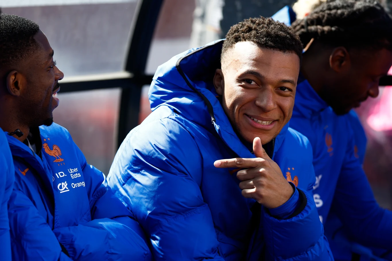 mężczyzna w niebieskiej kurtce sportowej siedzi wśród kolegów z drużyny, uśmiecha się szeroko i wskazuje na siebie palcem, wokół widoczni są współzawodnicy również ubrani w niebieskie kurtki