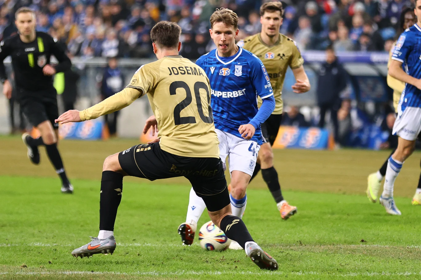 dwóch piłkarzy walczy o piłkę na zielonej murawie podczas meczu, jeden ubrany w niebieski strój, drugi w złoty z numerem 20 na plecach, w tle sędzia i pełne trybuny kibiców