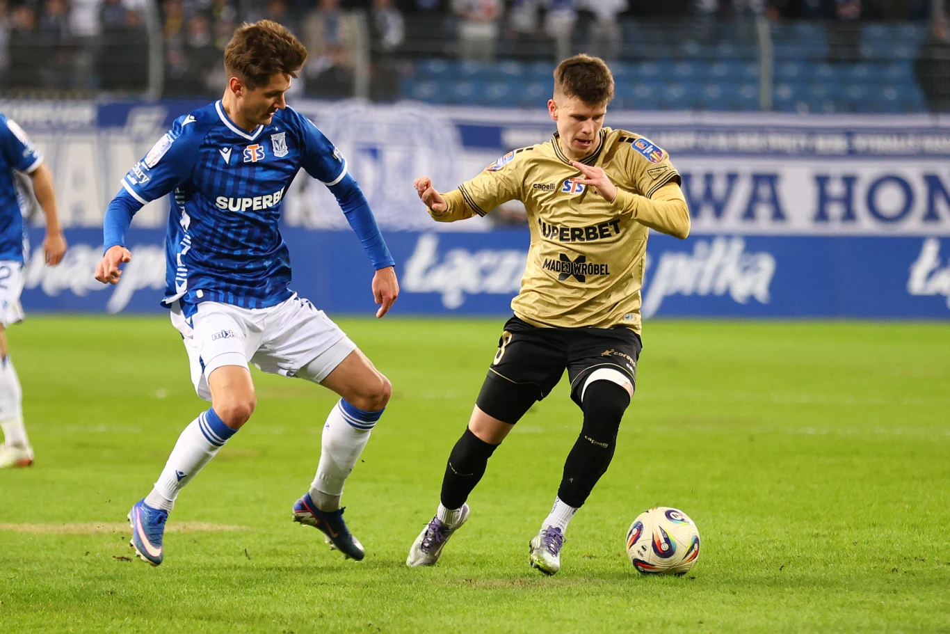 Dwóch piłkarzy rywalizuje o piłkę na murawie, jeden w stroju niebiesko-białym z logo sponsorów, drugi w złoto-czarnym, skoncentrowani podczas dynamicznej akcji meczu piłki nożnej.