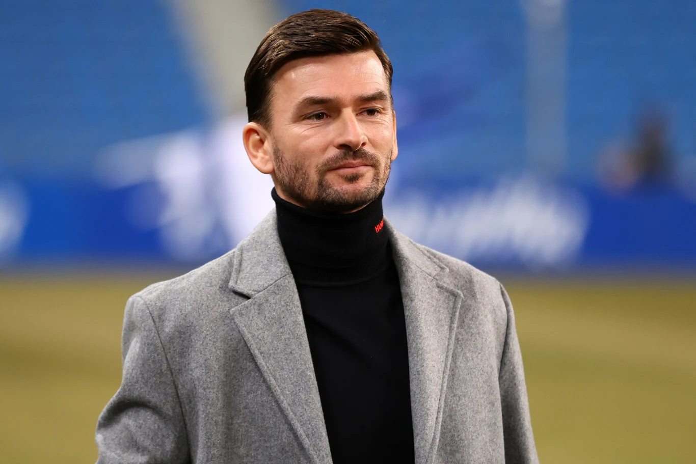 Mężczyzna z krótkimi, ciemnymi włosami ubrany w szary płaszcz oraz czarny golf stoi na tle niebieskich, rozmytych trybun stadionu.