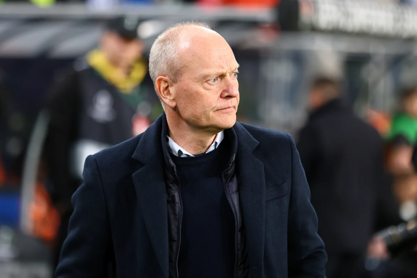łysy mężczyzna w eleganckim, ciemnym płaszczu i swetrze stoi na tle nieostrego stadionu, patrzy w bok z poważnym wyrazem twarzy