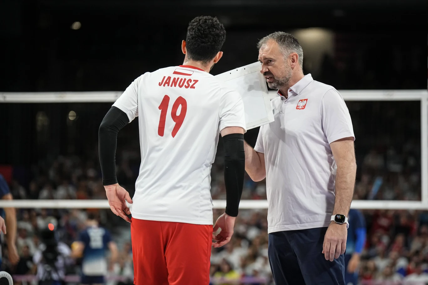 Trener polskiej reprezentacji siatkówki przekazuje taktyczne wskazówki zawodnikowi w białej koszulce z numerem 19 podczas meczu, trzymając przy twarzy kartkę z zapiskami, obaj skupieni na rozmowie.