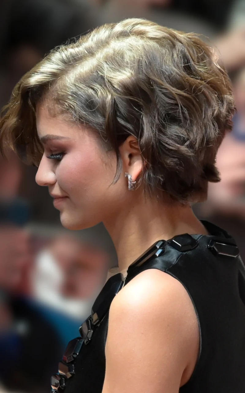 kobieta z krótkimi, falowanymi włosami w odcieniu ciemnego blondu, ubrana w elegancką, czarną sukienkę z ozdobnymi detalami na ramieniu, delikatny makijaż podkreślający rysy twarzy, subtelne kolczyki w uszach