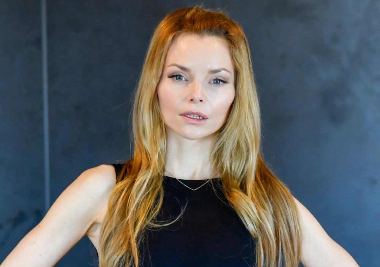 Izabella Miko Izabella Miko w czarnej, bezrękawnej bluzce, z długimi blond włosami, stoi na tle ciemnej ściany.