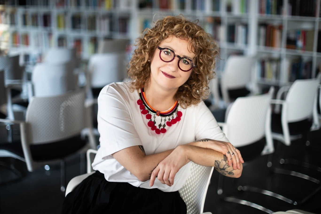 Uśmiechnięta kobieta z kręconymi blond włosami i okularami siedzi na białym krześle, opierając się na oparciu, w tle półki z książkami oraz rząd pustych, białych krzeseł.