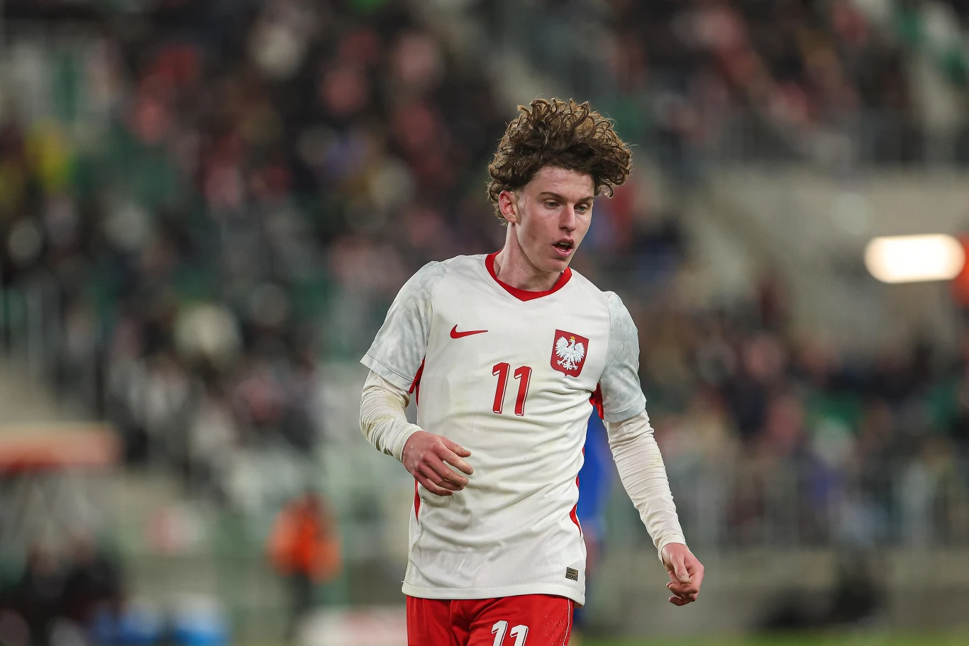 piłkarz w białej koszulce z numerem 11 i herbem Polski podczas meczu na stadionie, w tle rozmazana publiczność