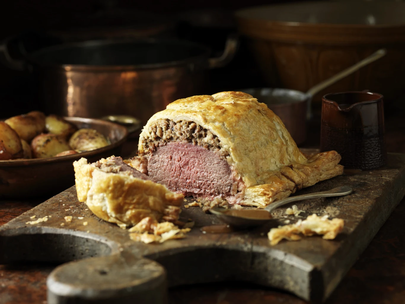 pokrojona pieczeń wołowa w cieście leży na drewnianej desce, obok znajdują się sztućce i kawałki kruchego ciasta; w tle widoczne są pieczone ziemniaki w garnku oraz niewielki dzbanek