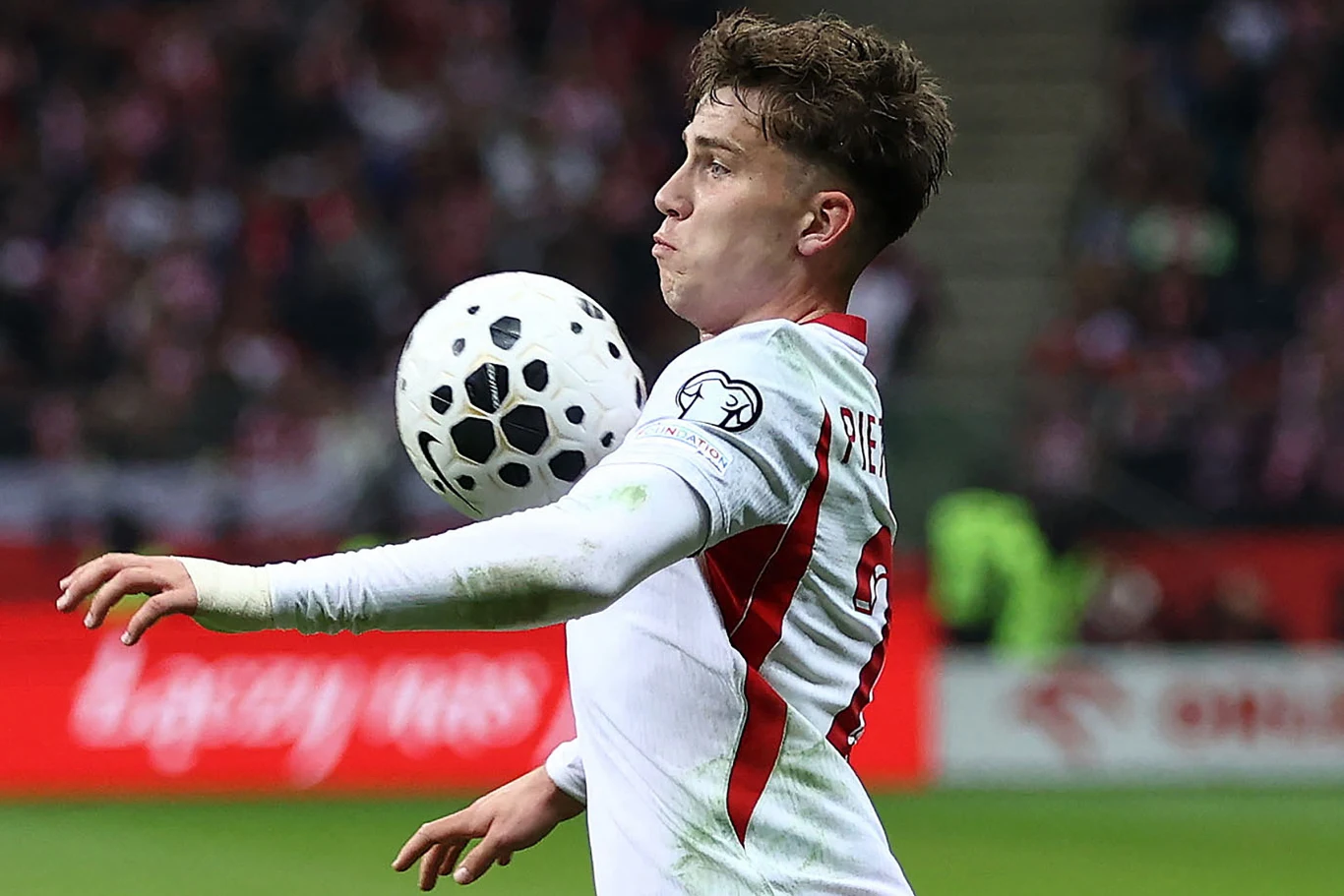 piłkarz w białej koszulce z czerwonym akcentem kontroluje piłkę klatką piersiową podczas meczu na stadionie, w tle rozmazana publiczność oraz baner reklamowy