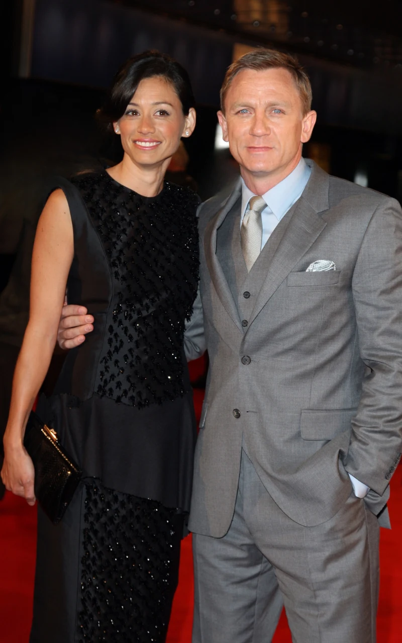 Satsuki Mitchell i Daniel Craig mężczyzna w szarym garniturze z krawatem i białą poszetką stoi obok kobiety ubranej w czarną elegancką sukienkę, trzymając ją za talię, oboje uśmiechnięci patrzą w stronę aparatu na tle czerwonego dywanu