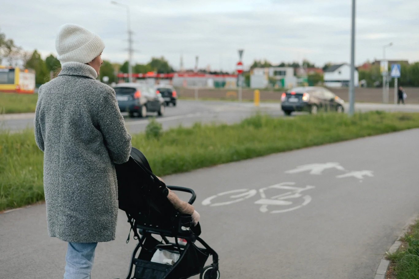 Osoba ubrana w szary płaszcz i jasną czapkę prowadzi wózek dziecięcy po ścieżce pieszo-rowerowej, obok ruchliwej ulicy z samochodami w tle.