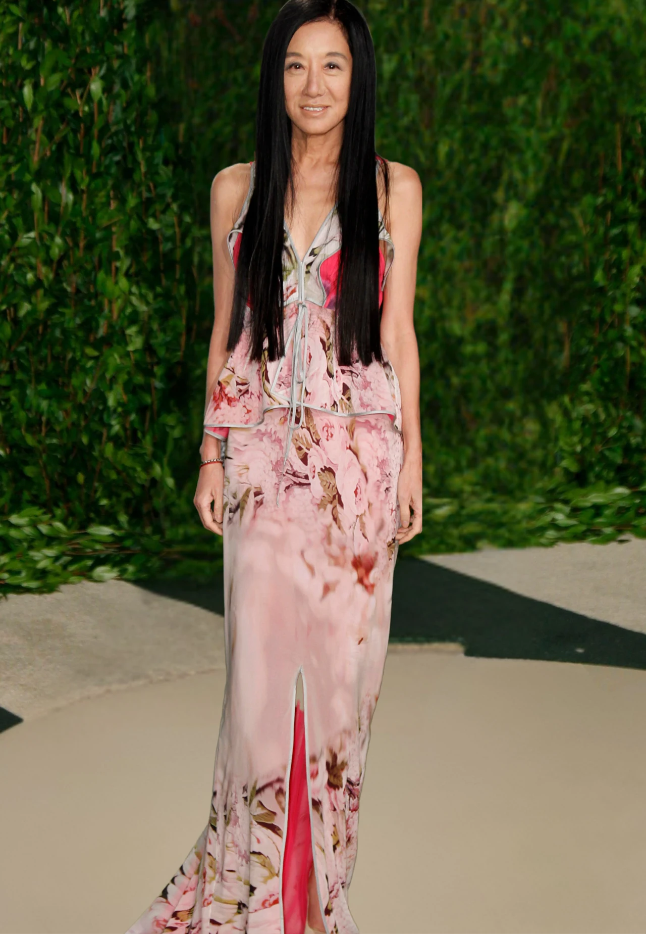 Vera Wang kobieta o długich, prostych, czarnych włosach ubrana w elegancką, długą suknię w kwiatowy wzór z rozcięciem na dole, stojąca na tle zielonej roślinności