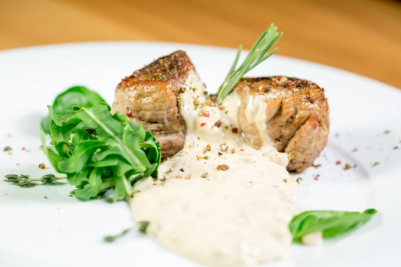 Soczysty stek polany kremowym sosem pieprzowym, udekorowany gałązką rozmarynu i podany z garścią świeżych liści rukoli oraz bazylią.
