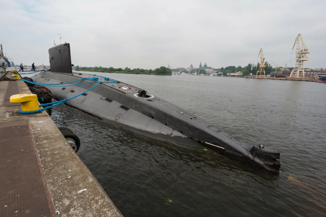 wojskowy okręt podwodny zacumowany przy betonowym nabrzeżu portowym, częściowo zanurzony w wodzie, z widocznymi linami cumowniczymi i żółtym odbojnikiem, w tle żurawie portowe oraz zabudowania stoczniowe