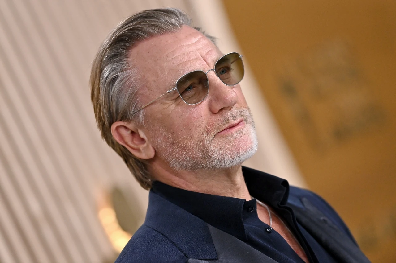 Daniel Craig mężczyzna w średnim wieku z siwą brodą noszący okulary przeciwsłoneczne oraz elegancki granatowy garnitur i koszulę, patrzy pewnie w bok na tle jasnych, geometrycznych ścian