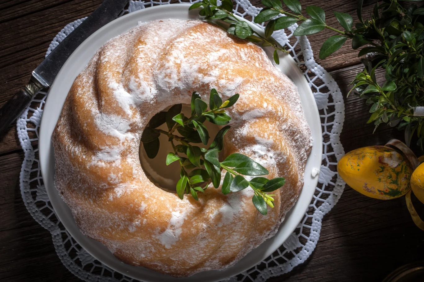 babka wielkanocna posypana cukrem pudrem, udekorowana gałązką bukszpanu, ustawiona na białym talerzu z koronkową serwetką na drewnianym stole obok ozdobnych żółtych pisanek