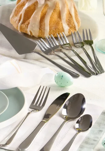 Sztućce starannie ułożone na białym obrusie obok pastelowych talerzy, połowa babki z polewą na talerzu oraz małe zielone jajko dekoracyjne pośrodku kompozycji.