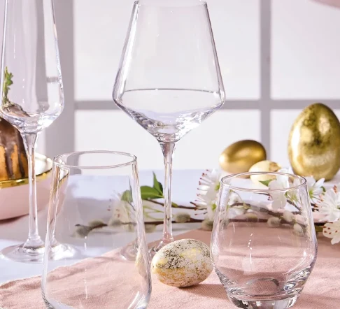 kilka różnych kieliszków i szklanek ustawionych na pastelowym obrusie, wśród ozdobnych jajek i kwiatowych gałązek, w tle złote wielkanocne jajka i ciasto