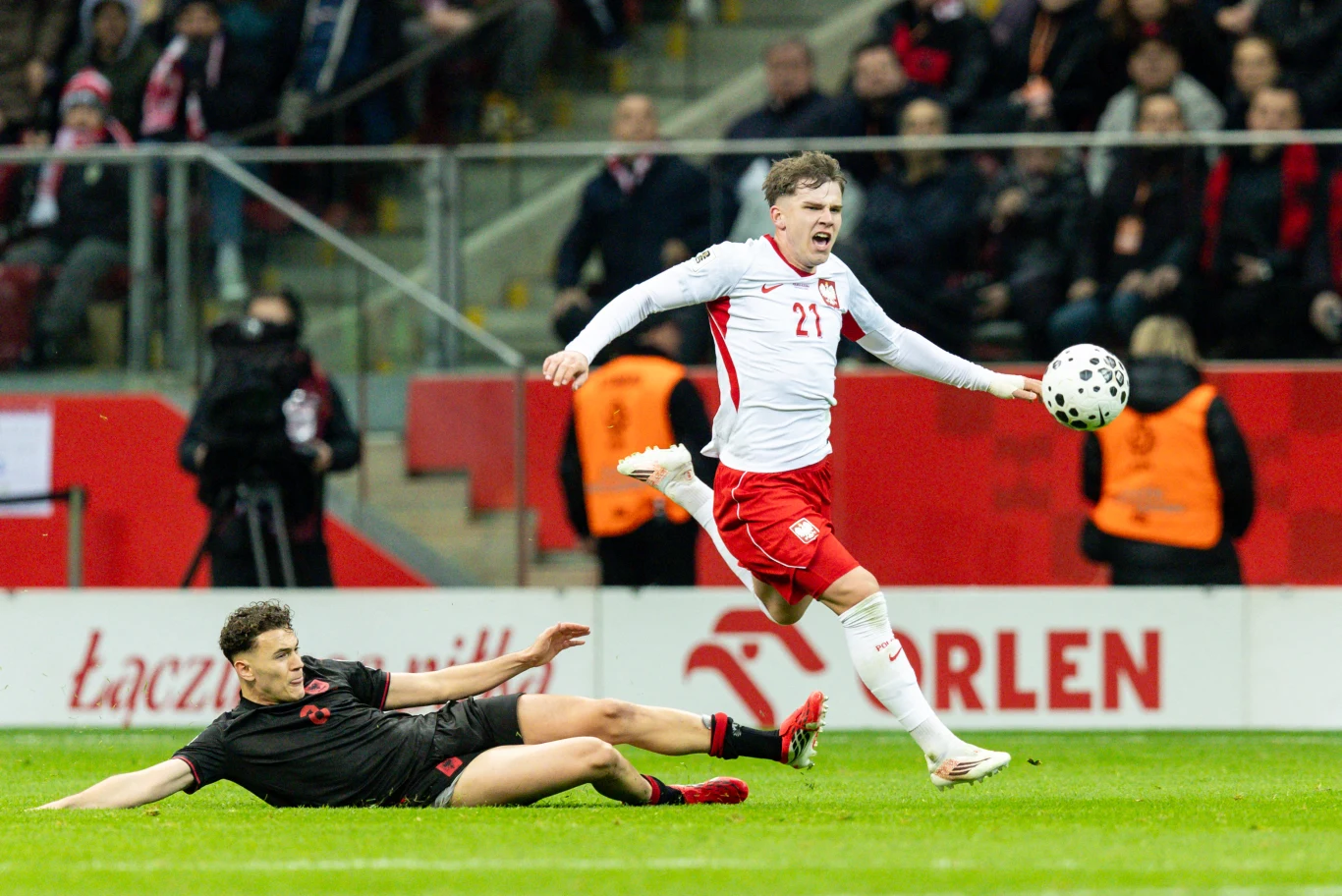 piłkarz w białej i czerwonej koszulce z numerem 21 biegnie z piłką, a zawodnik w czarnym stroju próbuje go powstrzymać ślizgiem na murawie na stadionie pełnym kibiców
