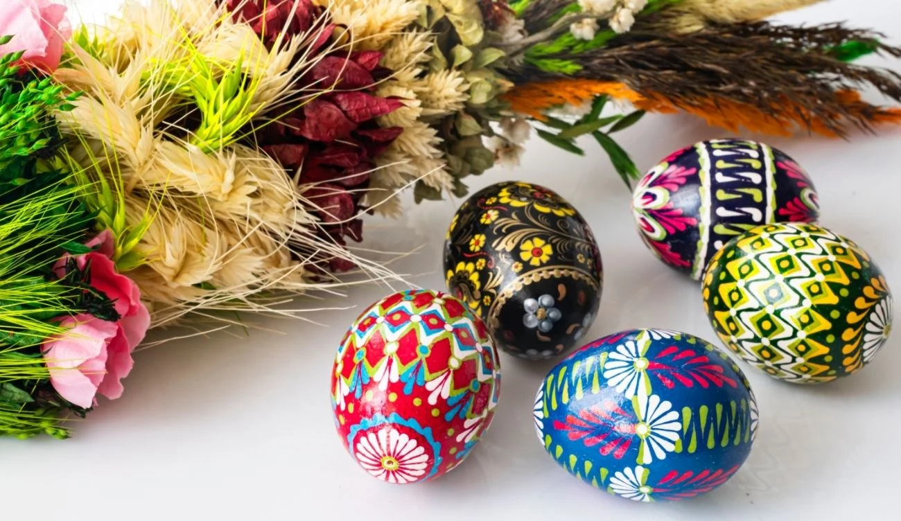 Jak dobrze znasz wielkanocne przysłowia? Pięć wielokolorowych pisanek i tradycyjna palma wielkanocna leżące na białym blacie.