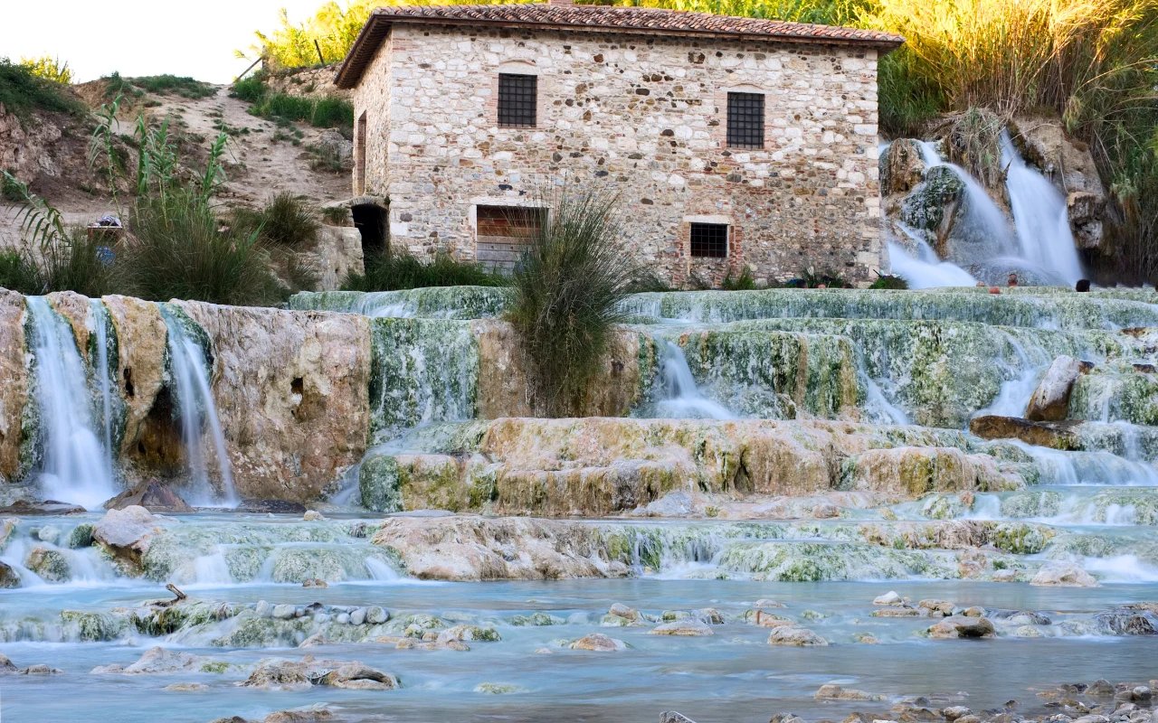 Cascate del Mulino w Saturnii – najpiękniejsze darmowe termy w Europie Kamienny budynek z brązowym dachem położony nad kaskadowymi wodospadami spływającymi po wapiennych tarasach, otoczony zielenią i naturalną roślinnością.