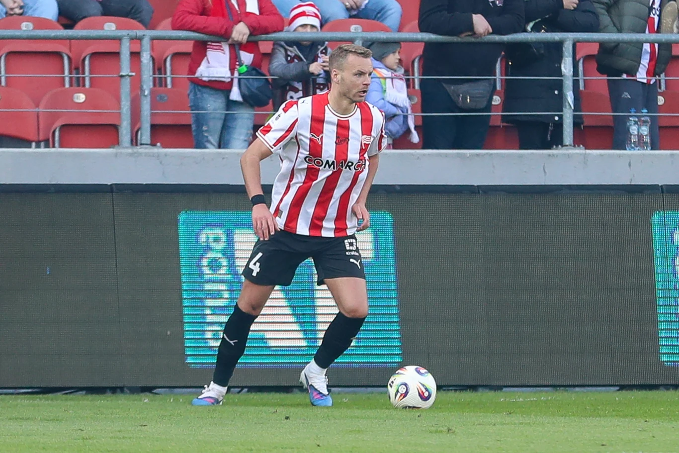 piłkarz w biało-czerwonej koszulce z numerem 4 kontroluje piłkę na murawie stadionu, w tle widoczna trybuna z kibicami