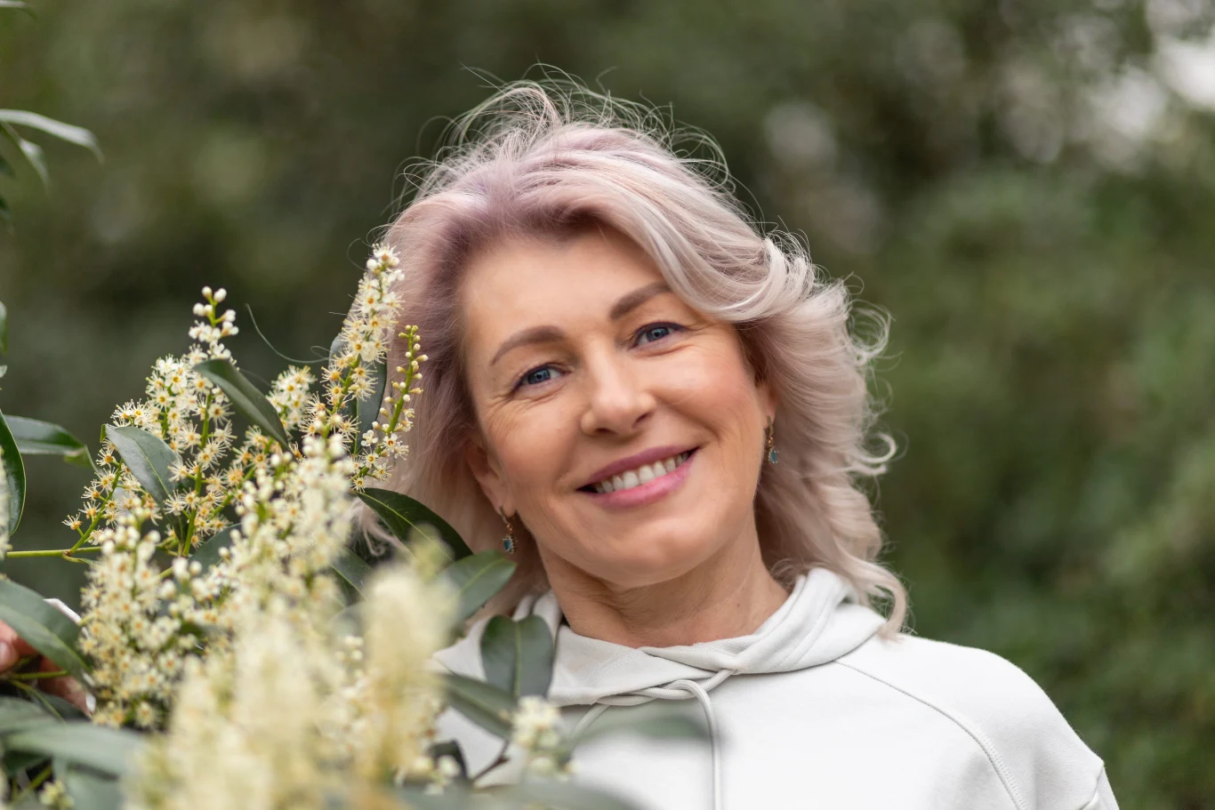 uśmiechnięta kobieta z jasnymi włosami w białej bluzie stoi wśród zieleni i gałązek z drobnymi białymi kwiatami