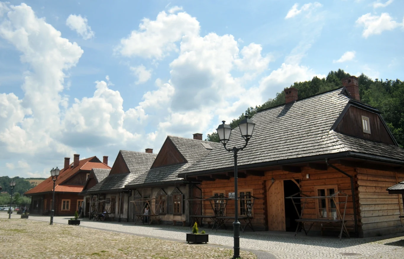 Miasteczko Galicyjskie to unikalna rekonstrukcja fragmentu małopolskiego miasteczka z przełomu XIX i XX wieku. drewniane, tradycyjne zabudowania ustawione w jednym rzędzie z brukowanym chodnikiem, latarnia uliczna na pierwszym planie, jasno oświetlone przez słońce, w tle zalesione wzgórza oraz fragment nieba z kłębiastymi chmurami