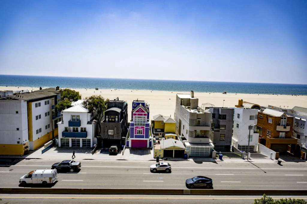 Szereg budynków frontem do szerokiej plaży i oceanu, w centrum wyróżnia się różowy dom w stylu inspirowanym bajką, przed domami biegnie ruchliwa droga z zaparkowanymi i przejeżdżającymi samochodami, w tle bezkresne morze i jasne, piaszczyste wybrzeże.