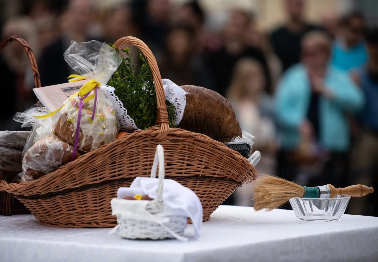 Święcenie pokarmów wiklinowy kosz wypełniony tradycyjnymi wielkanocnymi pokarmami, ozdobiony białą serwetką, obok mały koszyczek i kropidło z wodą święconą ustawione na białym obrusie.